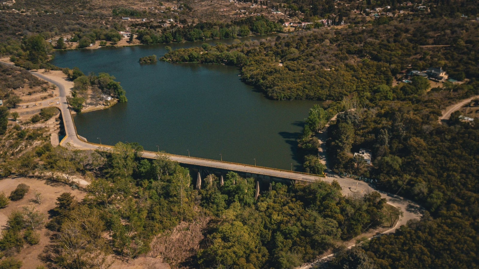 Fotografía aérea tomada con drone que muestra el muro de contención de la presa, el puente vehicular sobre ella y la extensión del lago rodeado de vegetación. Se aprecia la relación entre la obra civil y el cuerpo de agua estudiado.
