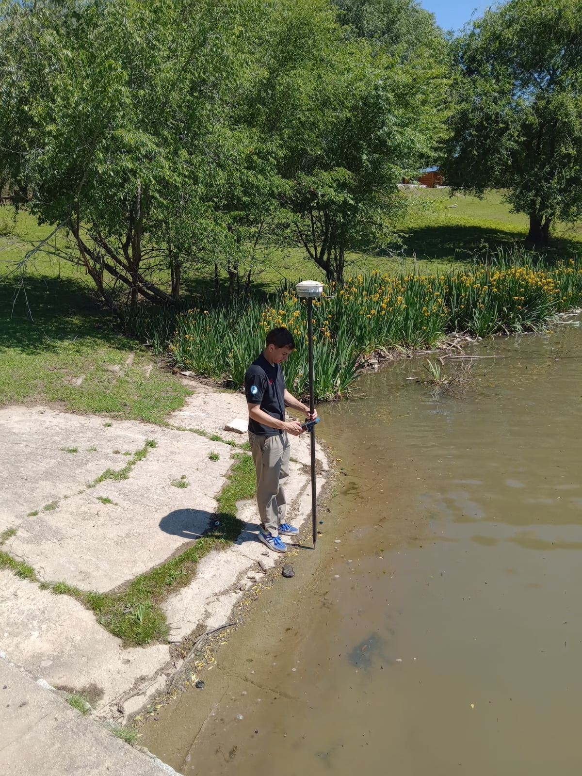 Agrimensor de pie en la orilla del lago operando un receptor GNSS/GPS montado en un bastón. El técnico está capturando datos de posicionamiento precisos para delimitar el perímetro del cuerpo de agua.